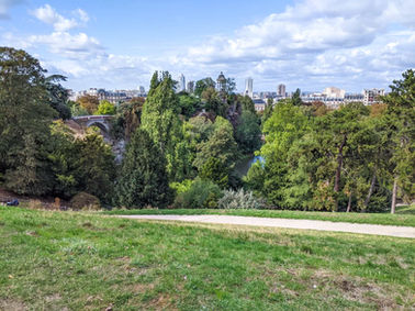 Green park overlook with trees, a stone bridge, river valley, and distant city buildings under a bright cloudy sky 11 unique things to do in Paris buttes chaumont park