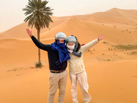 Two people in a desert with orange sand dunes and a palm tree. They wear helmets and scarves. Erg Chebbi dunes Sahara Desert Morocco