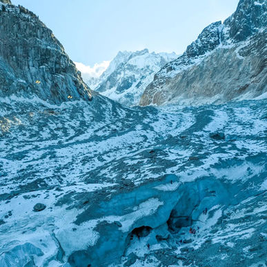 People explore a large blue glacier cave surrounded by snow-capped mountains. The sky is clear and bright blue, creating a serene atmosphere. Montenvers Mer de Glace Chamonix Mont Blanc France French Alps