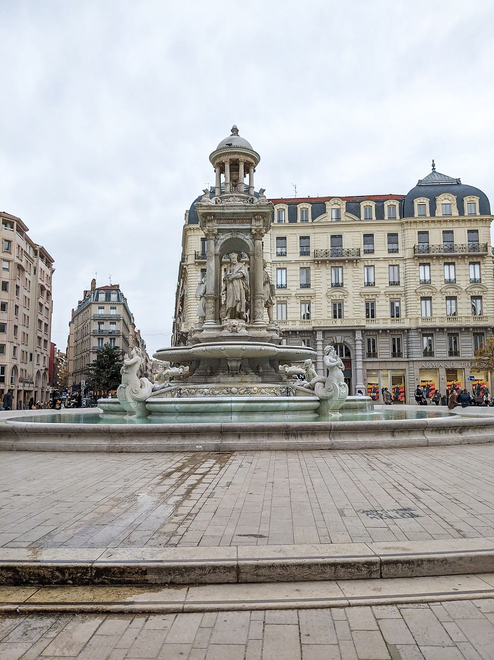 What to do in Lyon, France Historic fountain with sculptures in a European plaza, surrounded by classic buildings under a cloudy sky. People in the background.