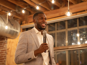 A Black entrepreneur in a light gray suit speaks confidently into a microphone while pitching at an event with warm string lights overhead.