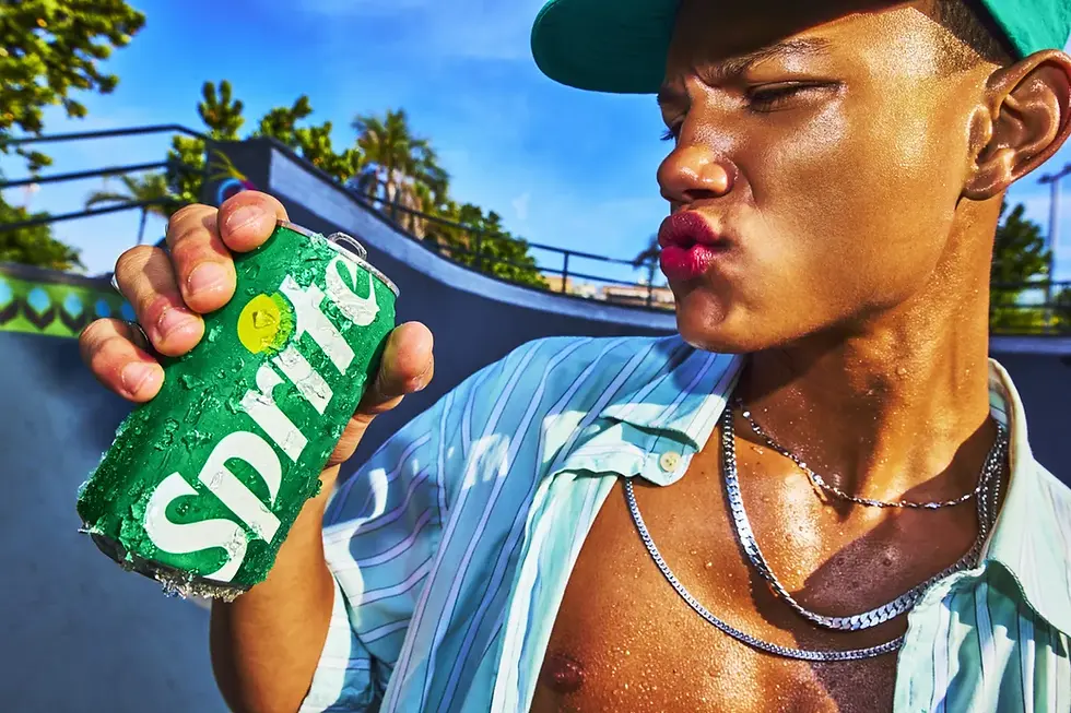 Close-up of a sweaty young person at a skate park holding a cold green Sprite can covered in water droplets, with palm trees and a blue sky in the background.