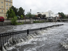"Floodwaters from a swollen river overflow a black metal fence along Main Street Plaza in a downtown area, following historic August 2025 flooding in Southeast Wisconsin."