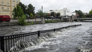 "Floodwaters from a swollen river overflow a black metal fence along Main Street Plaza in a downtown area, following historic August 2025 flooding in Southeast Wisconsin."