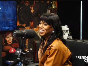 Kerry Washington smiles while speaking into a microphone during her guest appearance on The Breakfast Club radio show, seated in a studio with branded signage and cultural memorabilia in the background.