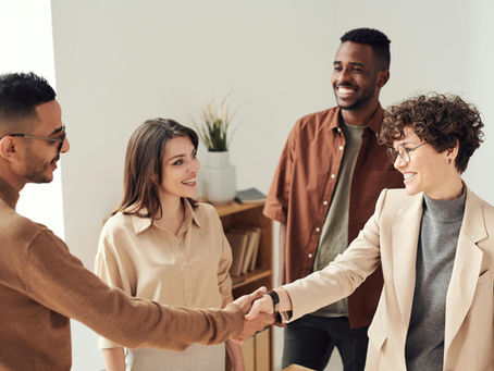 Four people in an office setting, smiling and shaking hands. Neutral-toned clothing and potted plant in the background convey a welcoming mood.