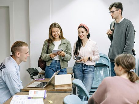 Students in a classroom gather around a table with a laptop. Two are smiling, holding papers. Others sit, creating a relaxed, studious mood.