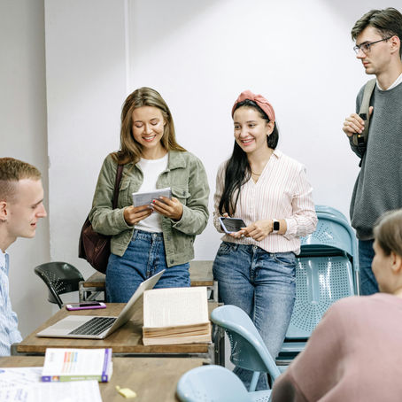 Students in a classroom gather around a table with a laptop. Two are smiling, holding papers. Others sit, creating a relaxed, studious mood.