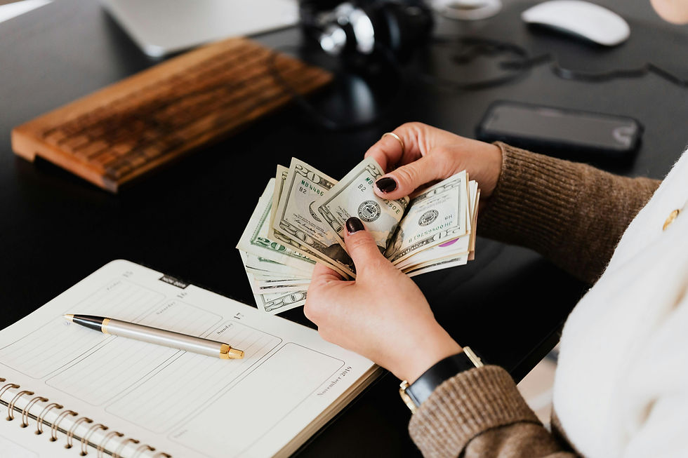 Hands counting US dollars at a dark desk with a wooden keyboard and planner. A pen lies on an open notebook, tablet in the background.