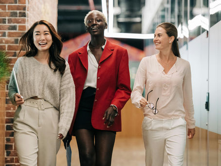 Three women walk down a hallway smiling. One wears a red blazer, another a gray sweater, and the third a white blouse, creating a cheerful mood.