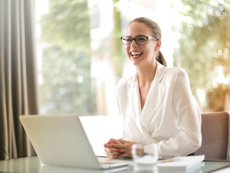 Smiling woman in glasses and white blazer sits at a desk with a laptop. Bright, blurred background creates a cheerful, professional mood.