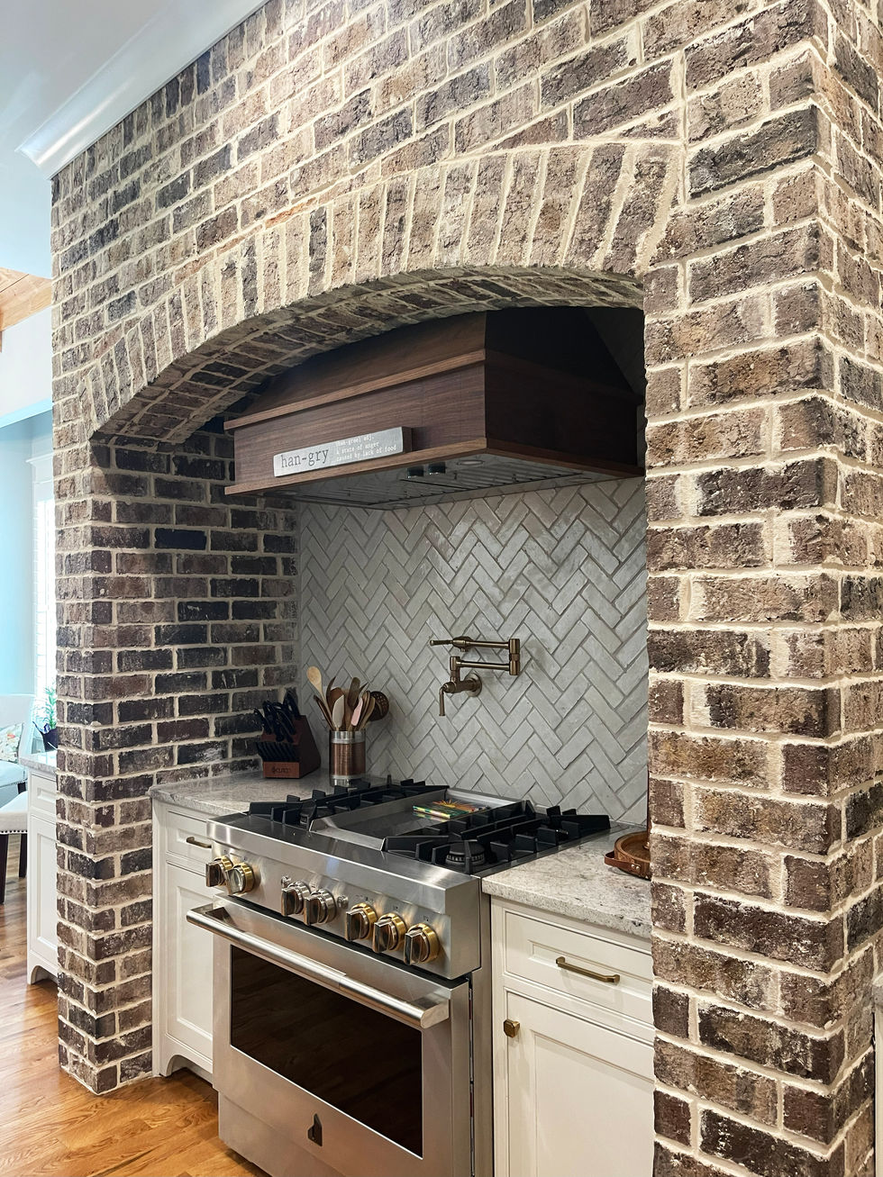 Custom kitchen design in Smyrna, GA home featuring brick arch surround, professional gas range, herringbone tile backsplash, and custom wood vent hood. 