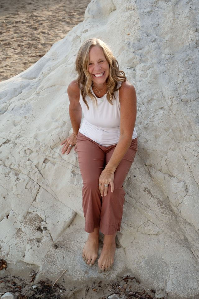 Kim Love smiling, sitting on large beach rocks outdoors.