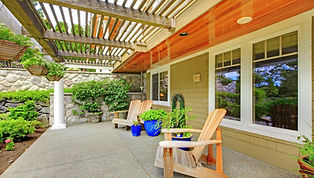 Photo of a backyard with a concrete pad under a pergola attached to the house, adorned with potted plants and Muskoka chairs.