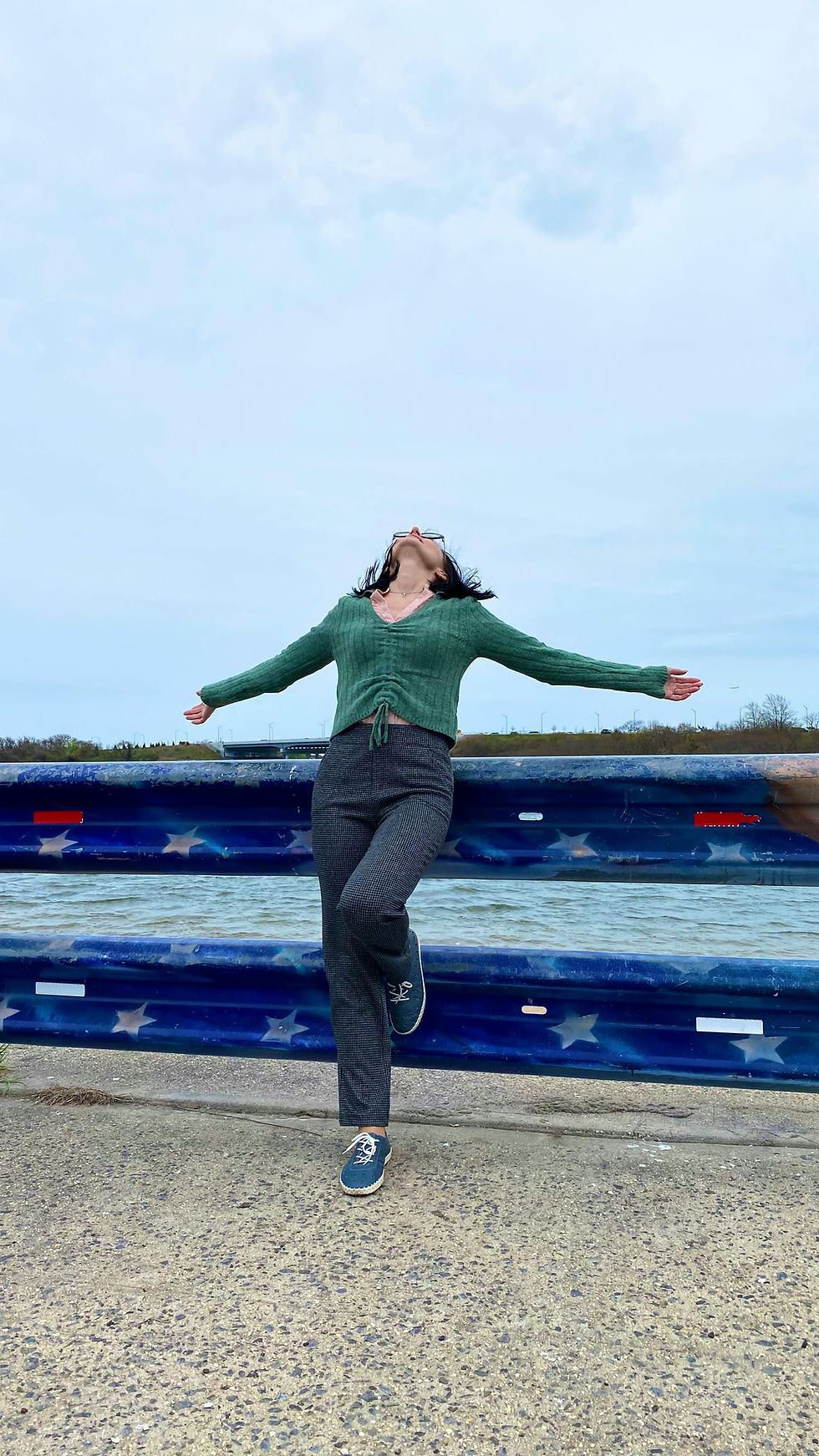Woman in green sweater and gray pants stretches arms joyfully by a railing with a star pattern, against a cloudy sky and water backdrop.