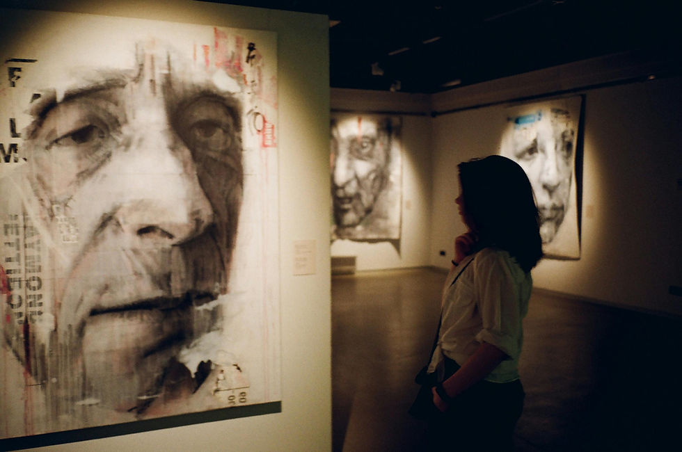 Woman observing large portrait paintings in dimly lit gallery. The artworks depict somber faces in black, white, and subtle red tones.