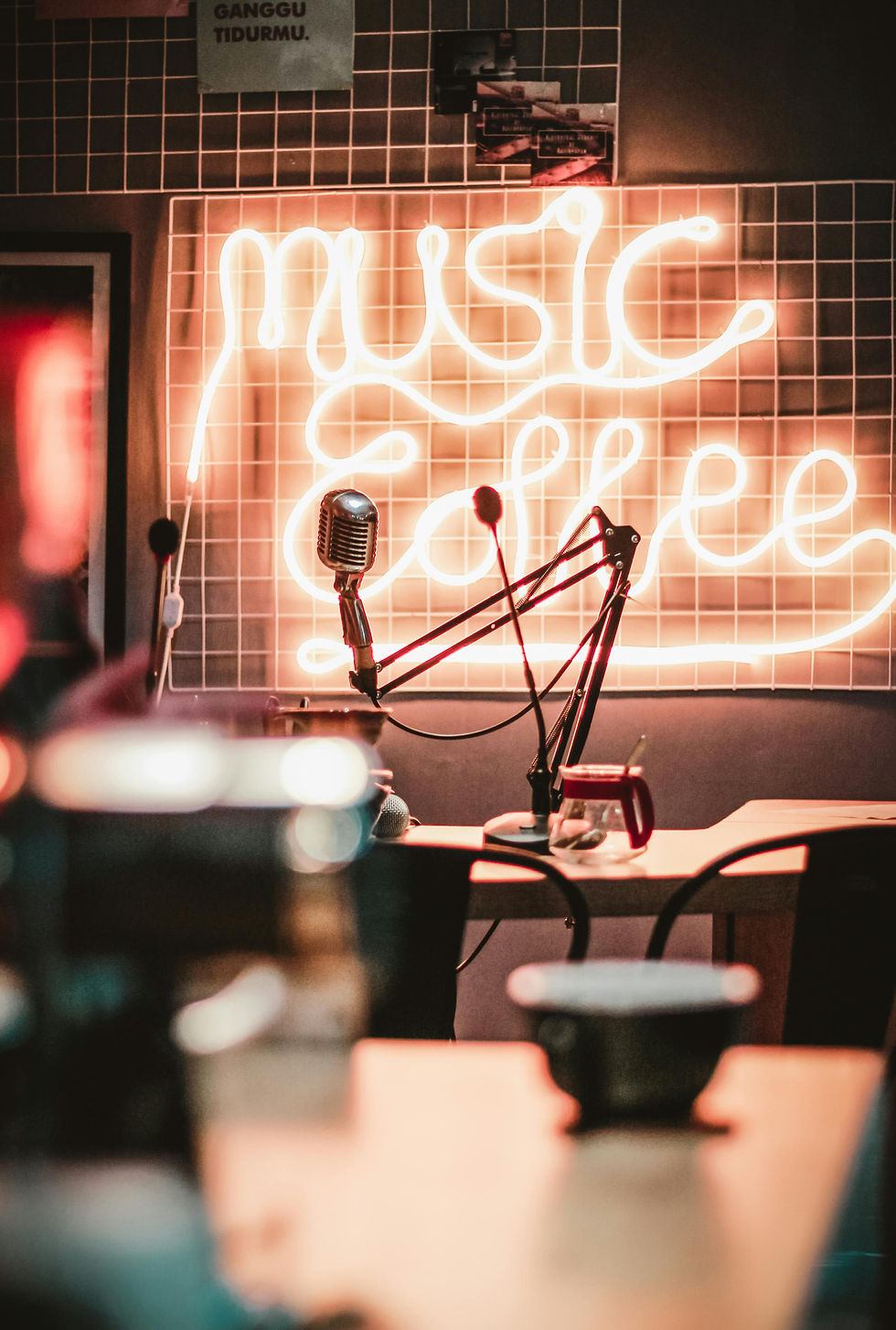 Vintage microphone and arm on a table, with "music coffee" neon sign in the background. Warm, cozy atmosphere with blurred items.