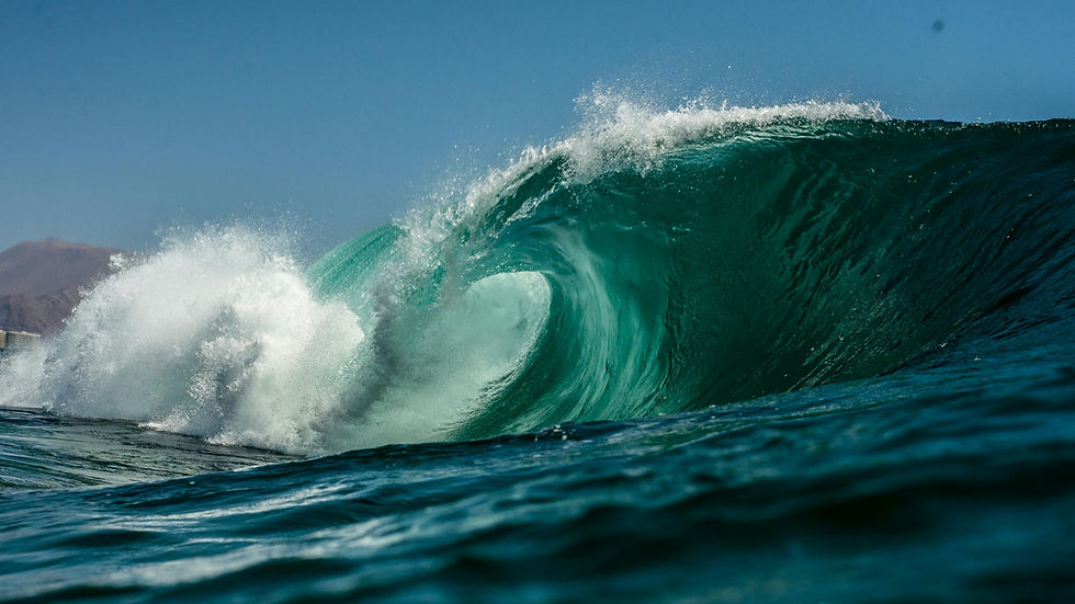 A powerful turquoise ocean wave crashes under a clear blue sky. Mountains are faintly visible in the background. Dynamic and energetic mood.