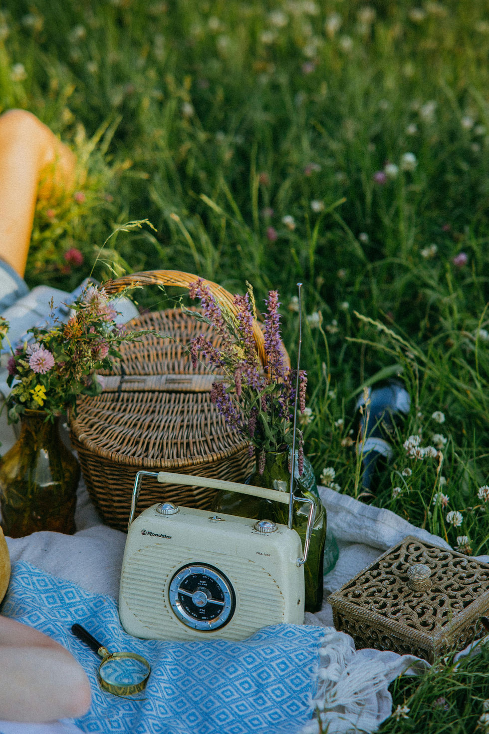 Vintage radio and wicker basket with flowers on a blue blanket in a grassy field. Relaxed picnic vibe with a magnifying glass nearby.