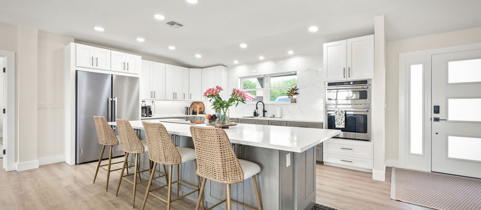 Modern kitchen with white cabinets, island with wicker chairs, gray floor, and stainless appliances. Bright ambiance with pink flowers.