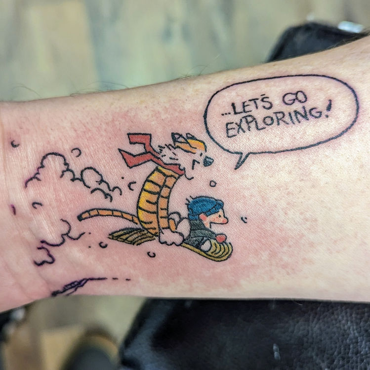 Calvin and Hobbes tattoo on an arm, showing Calvin riding Hobbes on a sled with the words '...LET'S GO EXPLORING!' in a speech bubble