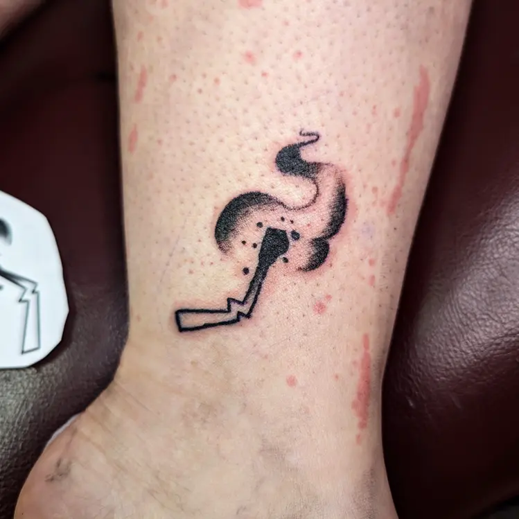 Black and white tattoo of a lit match with smoke on a person's ankle