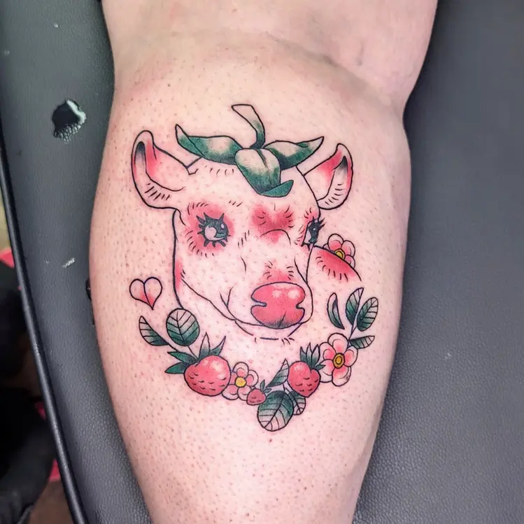 A pink cow with a strawberry on its head surrounded by flowers and strawberries, a tattoo on a person's leg.