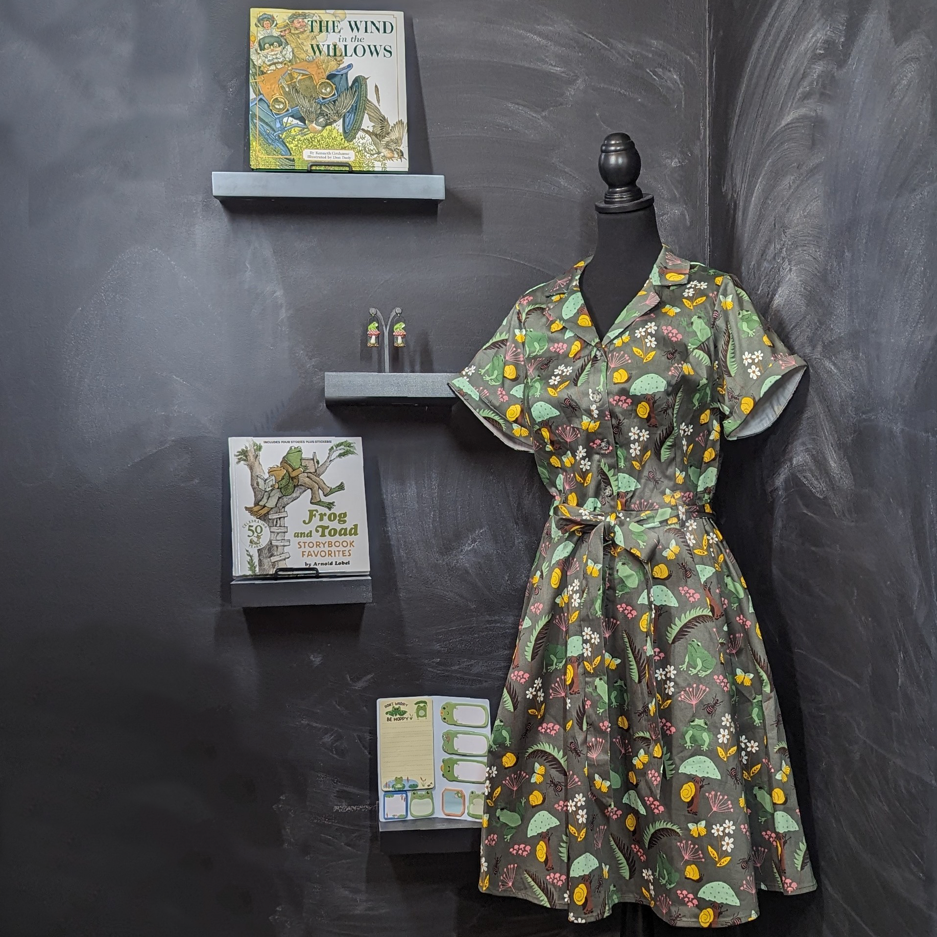 Picture of frog print dress, clay polymer frog statement earrings, The Wind in the Willow illustrated chapter book, The Frog and Toad Storybook Favorites picture book, and frog themed sticky notes. 