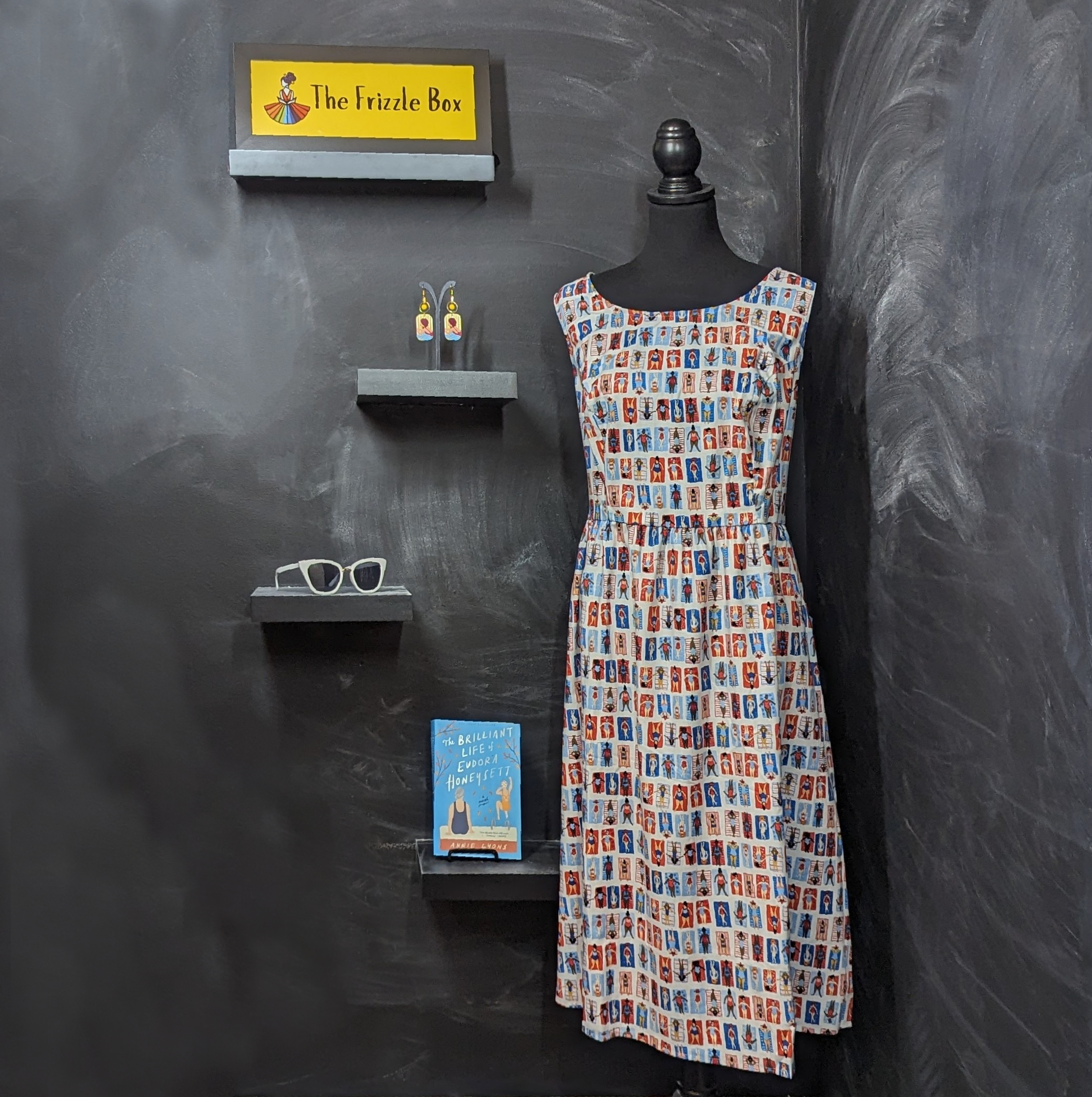 Picture of sunbather print dress, sunbather statement earrings, white cat-eye sunglasses, the adult summer read The Brilliant Life of Eudora Honeysett, and the Frizzle Box logo. 