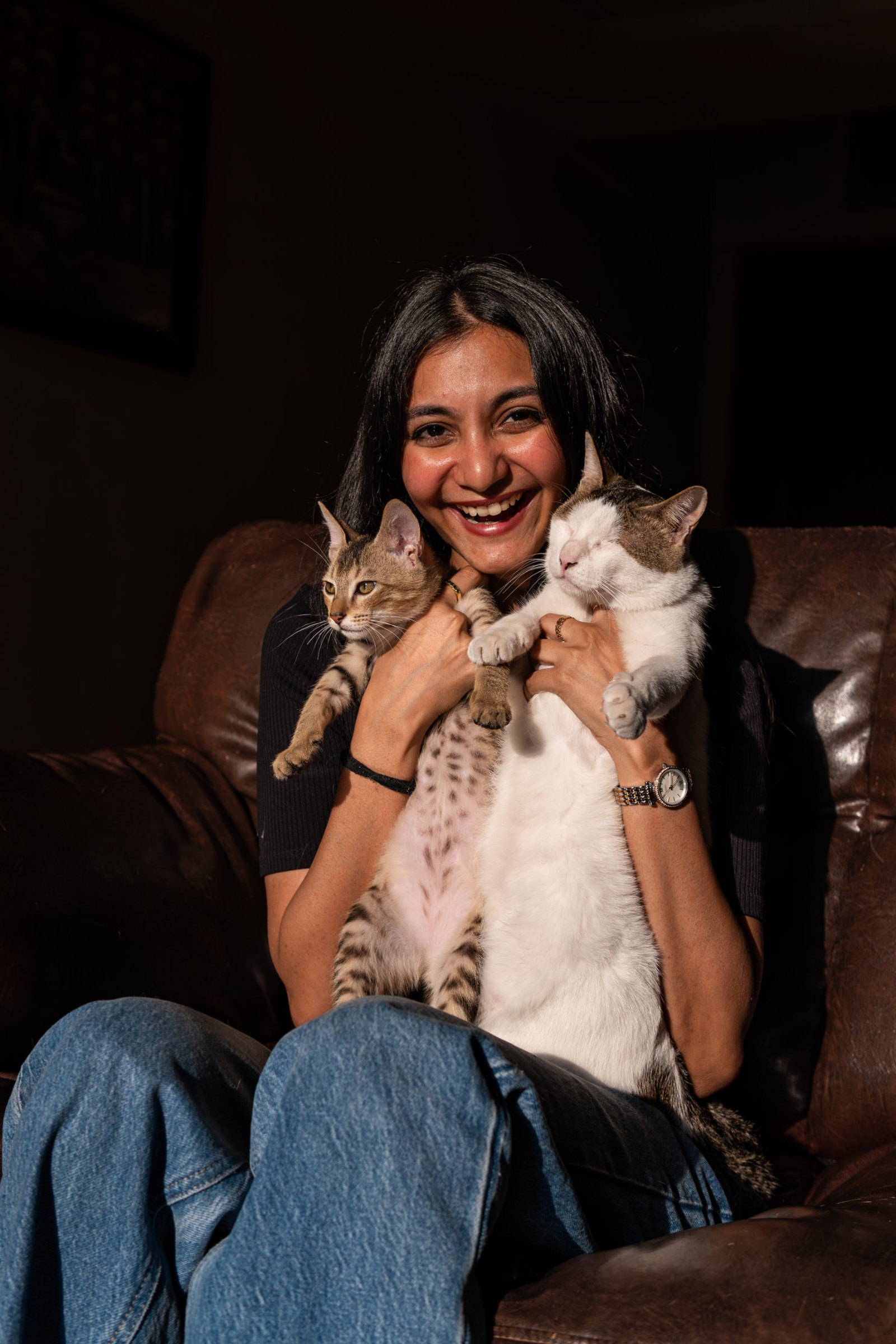 Nivedita laughs as she holds two cats, a tabby kitten and Loki, on a leather sofa