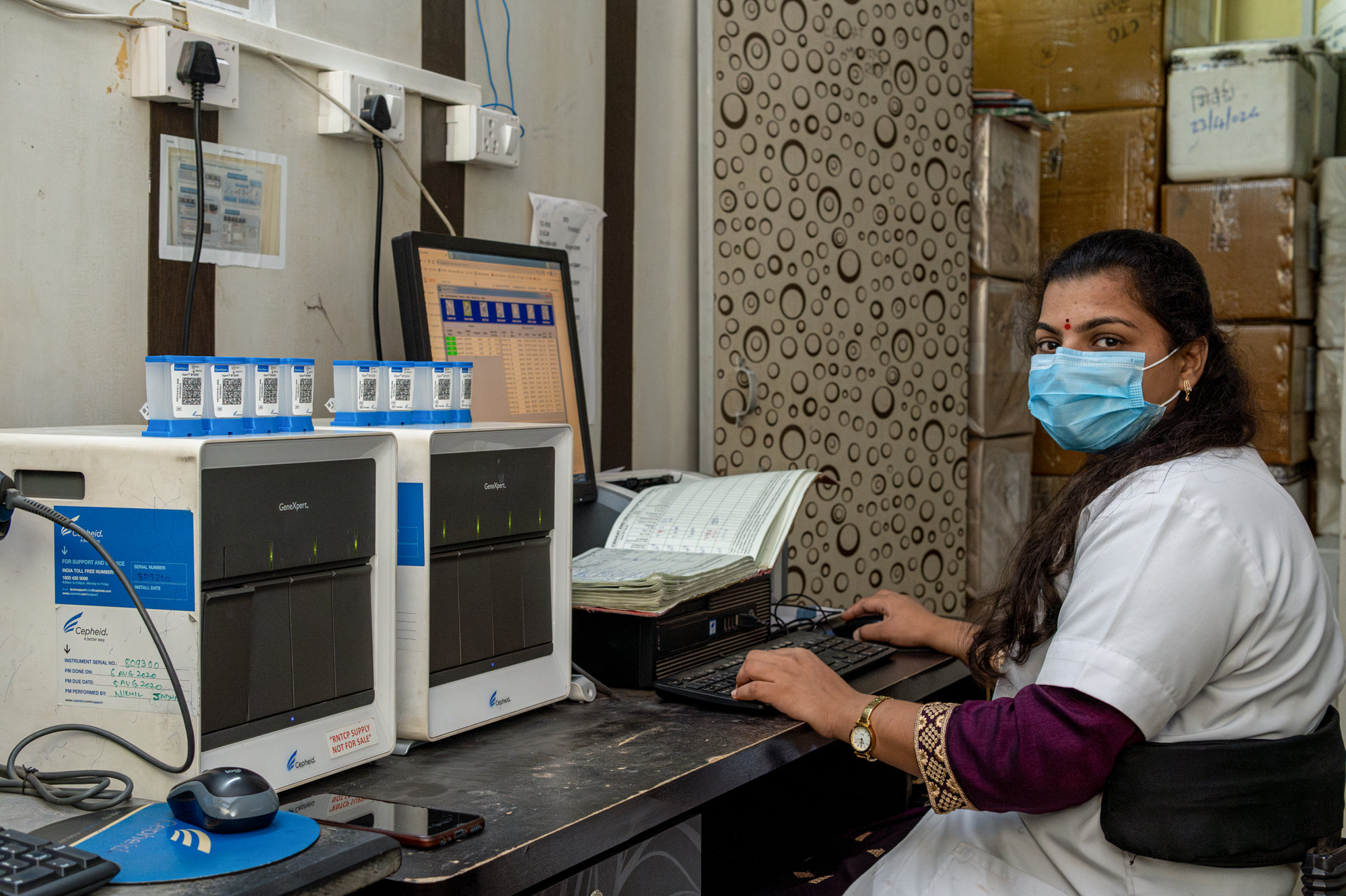 A lab technician in a mask works at a computer beside two GeneXpert testing machines