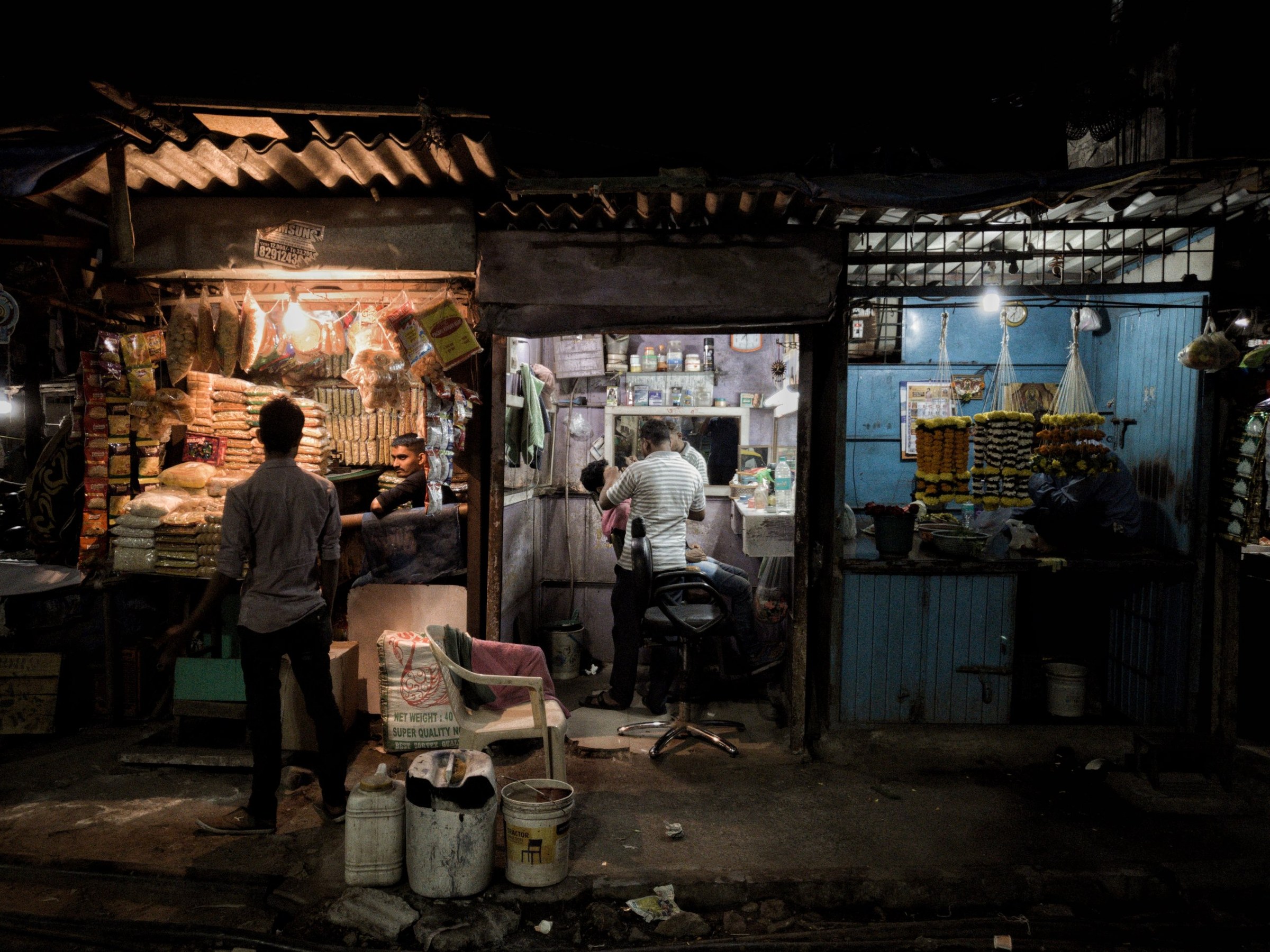 A row of small shops at night: a snack stall, a barber at work and a flower stall hung with marigold garlands