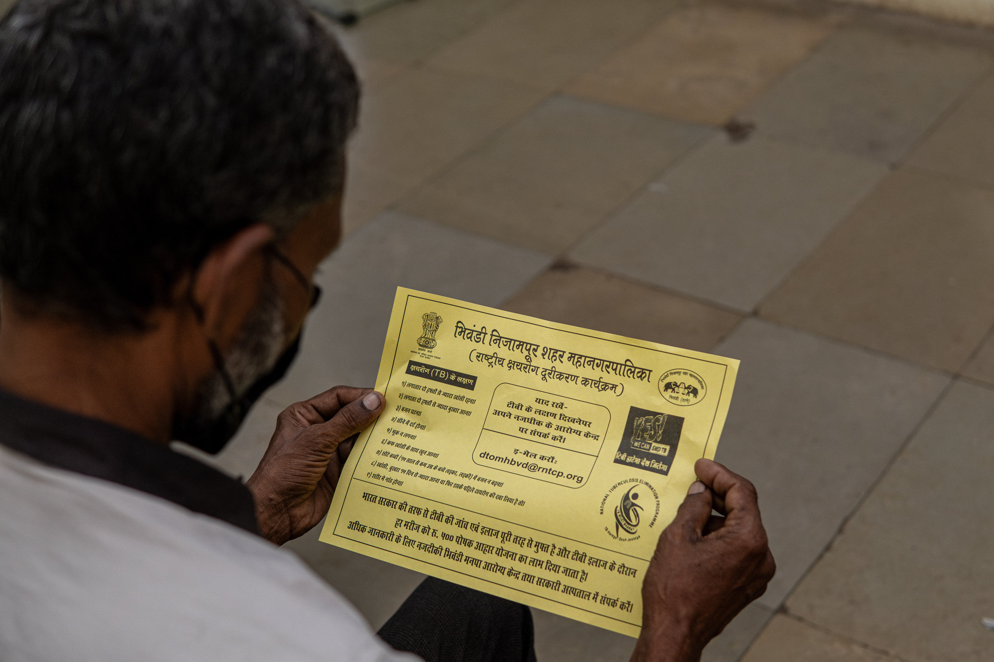 A man reads a yellow municipal leaflet in Hindi listing the symptoms of tuberculosis