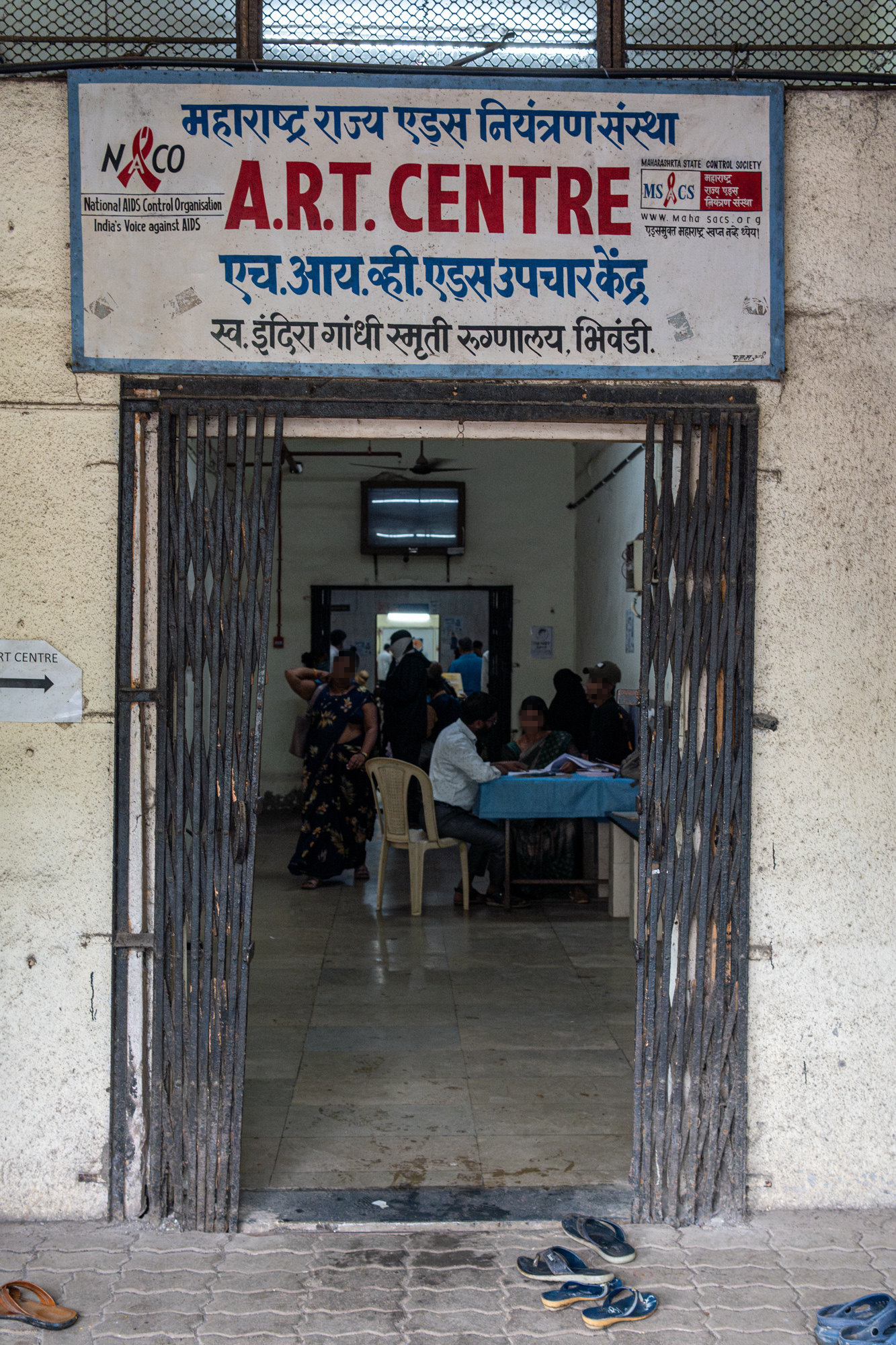 The barred doorway of the ART centre at a hospital in Bhiwandi, with staff and patients inside and sandals left at the entrance