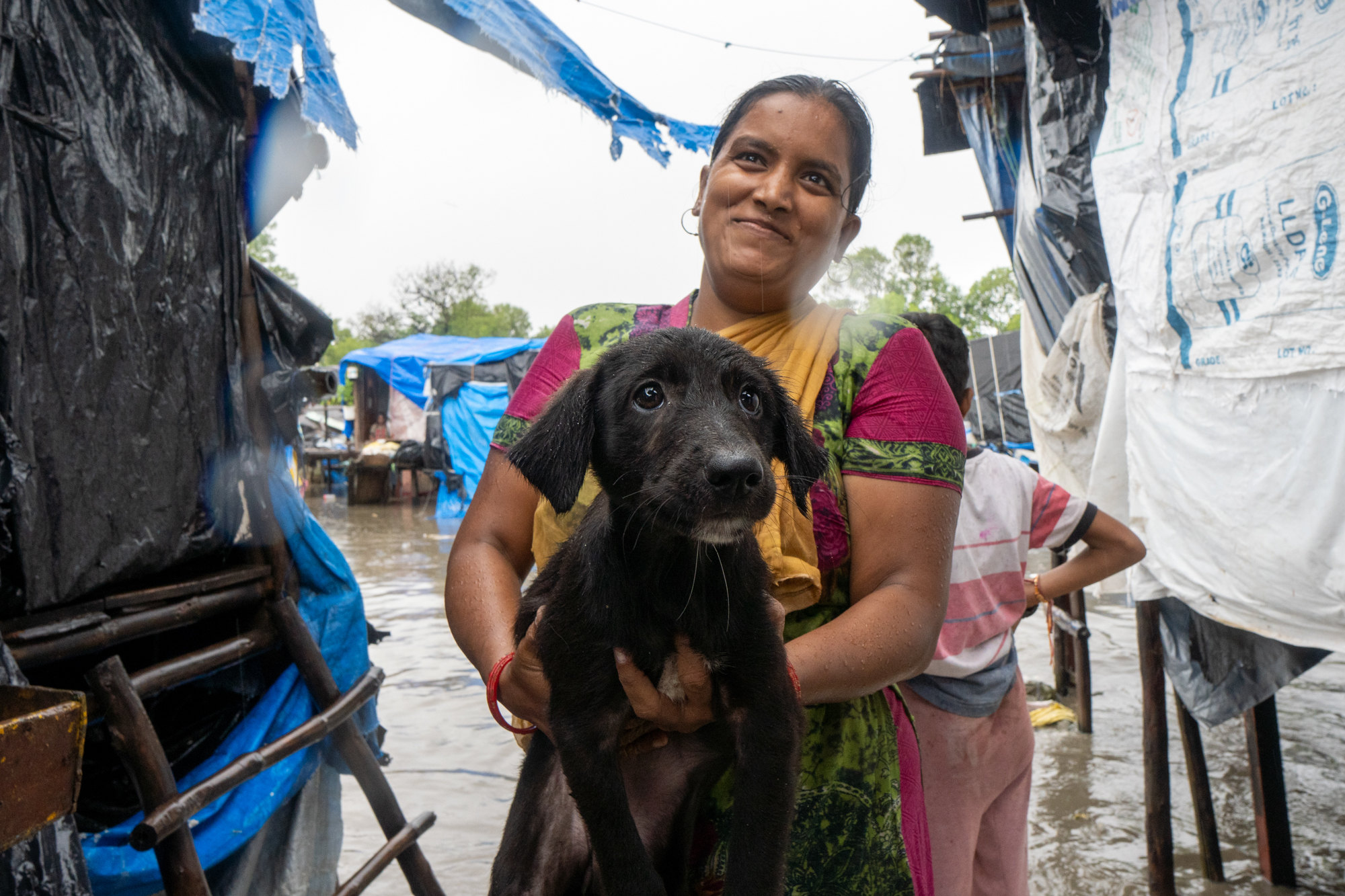 A smiling woman holds a wet black puppy in a flooded settlement
