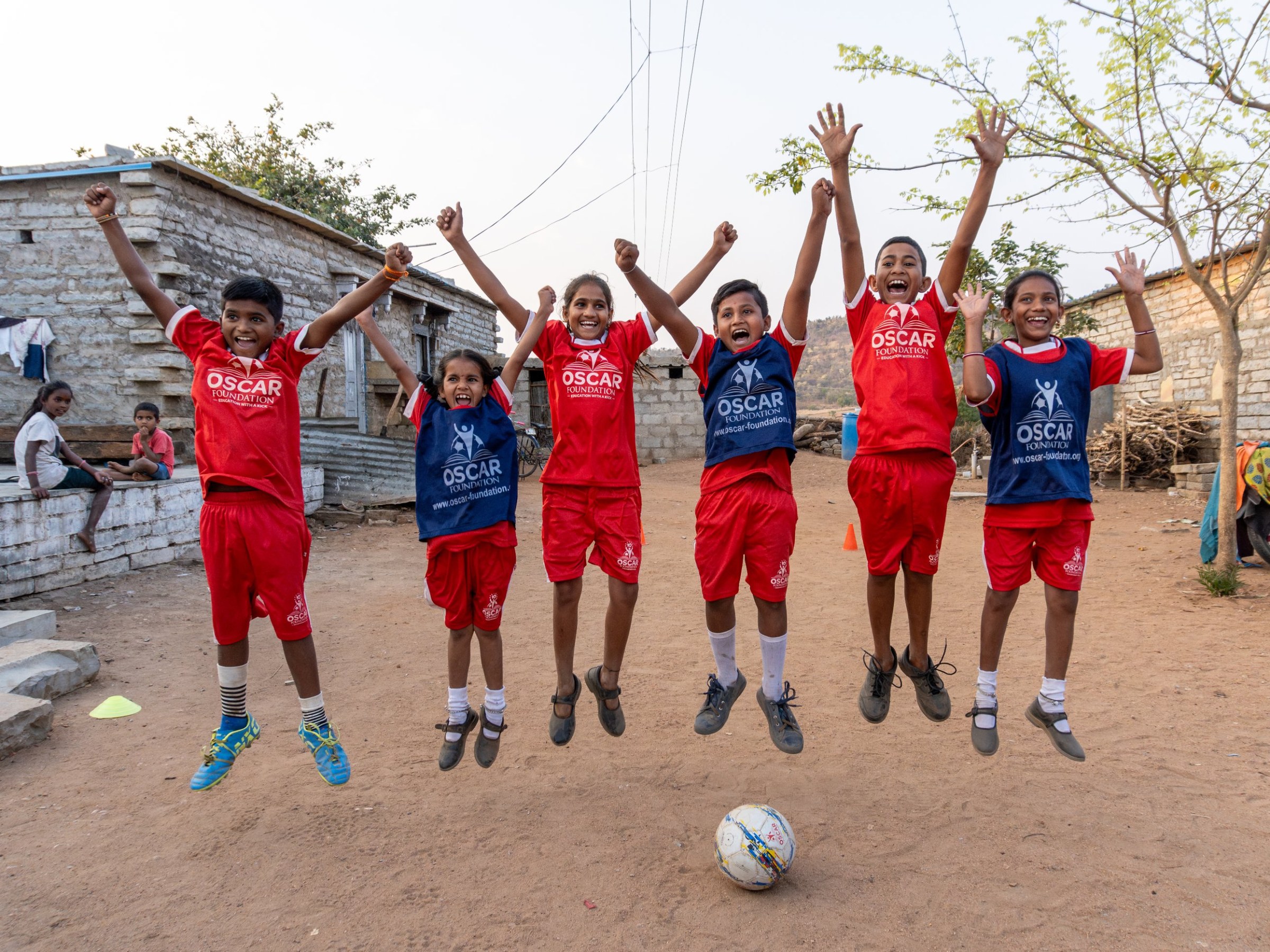 Six children in OSCAR Foundation football kits jumping in the air with arms raised, a ball at their feet