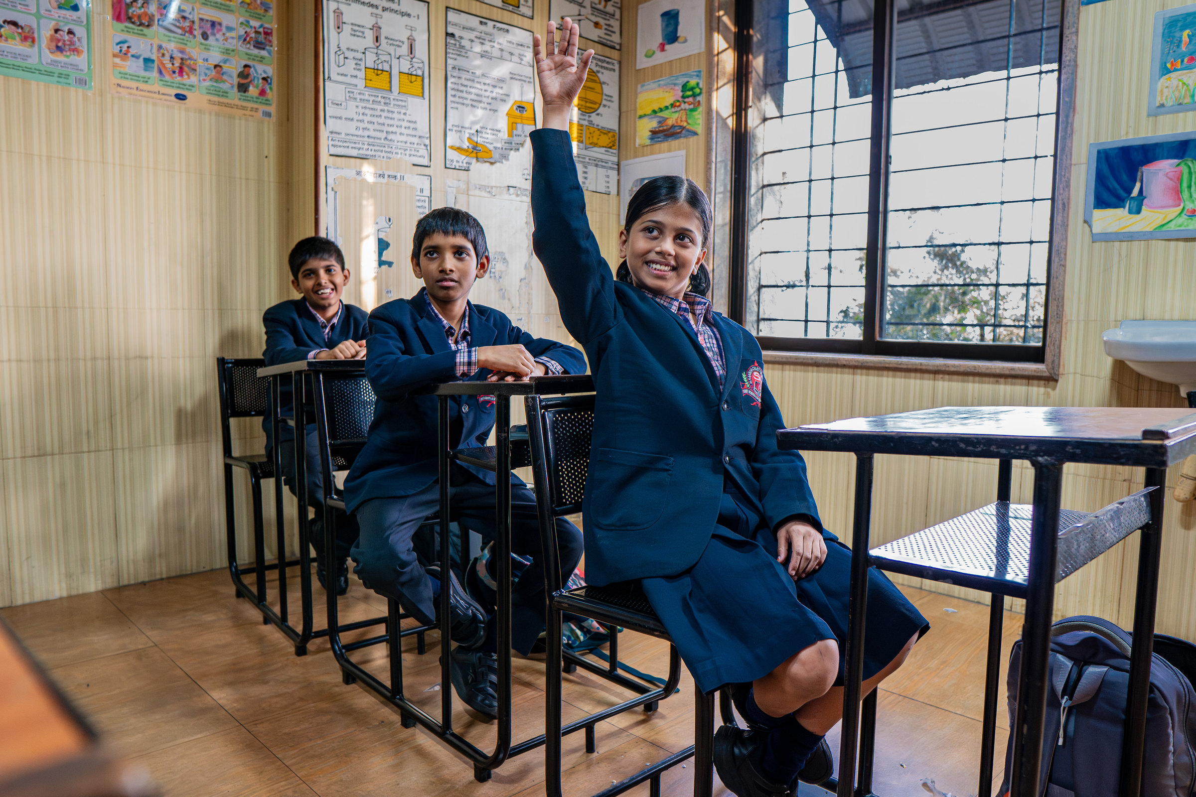 A girl smiles and raises her hand in a classroom, two classmates seated beside her
