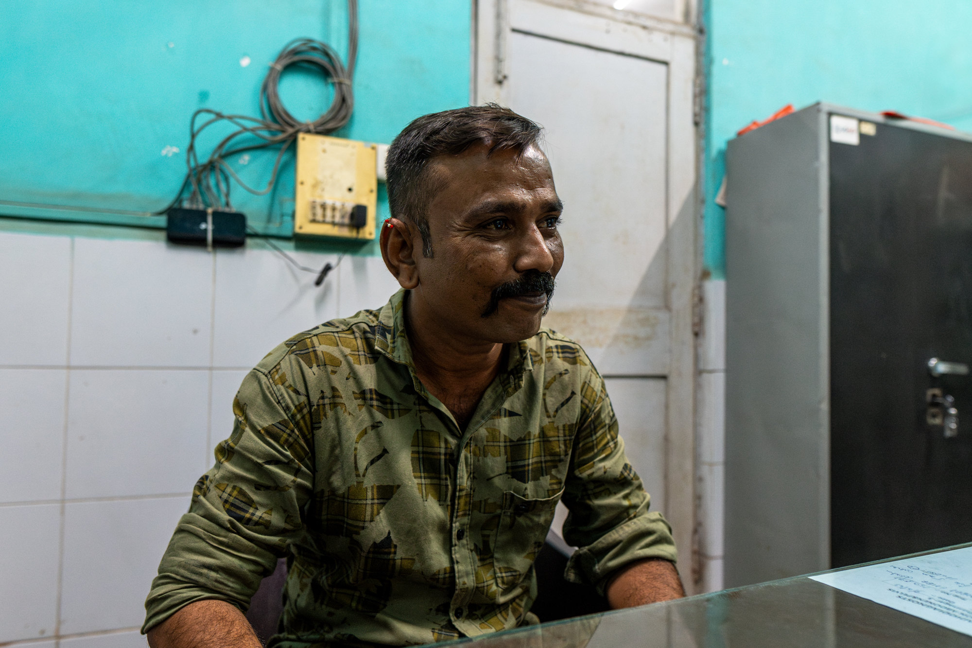A smiling man in a patterned shirt sits at a desk in a turquoise-walled office