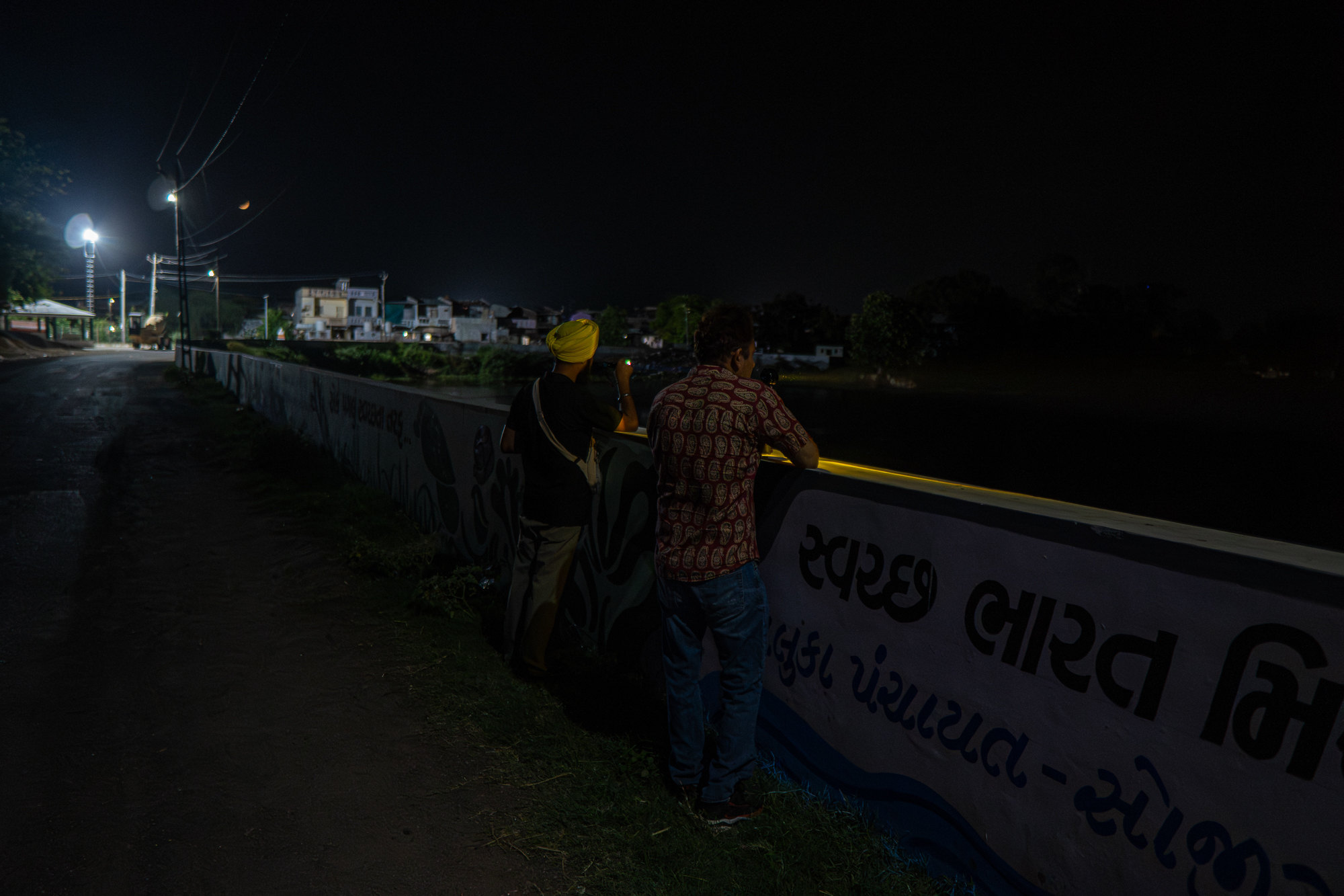 Two men lean over a lakeside wall at night, scanning the water with torches