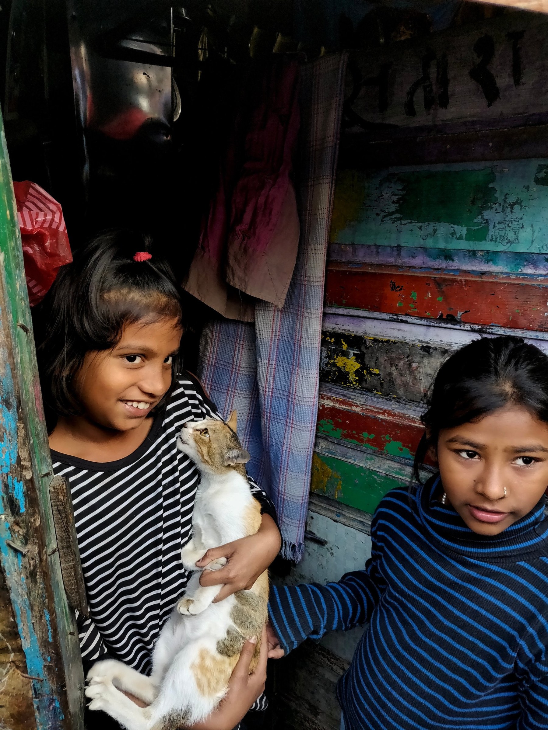Two girls in a doorway in Thane, one cradling a cat who gazes up at her