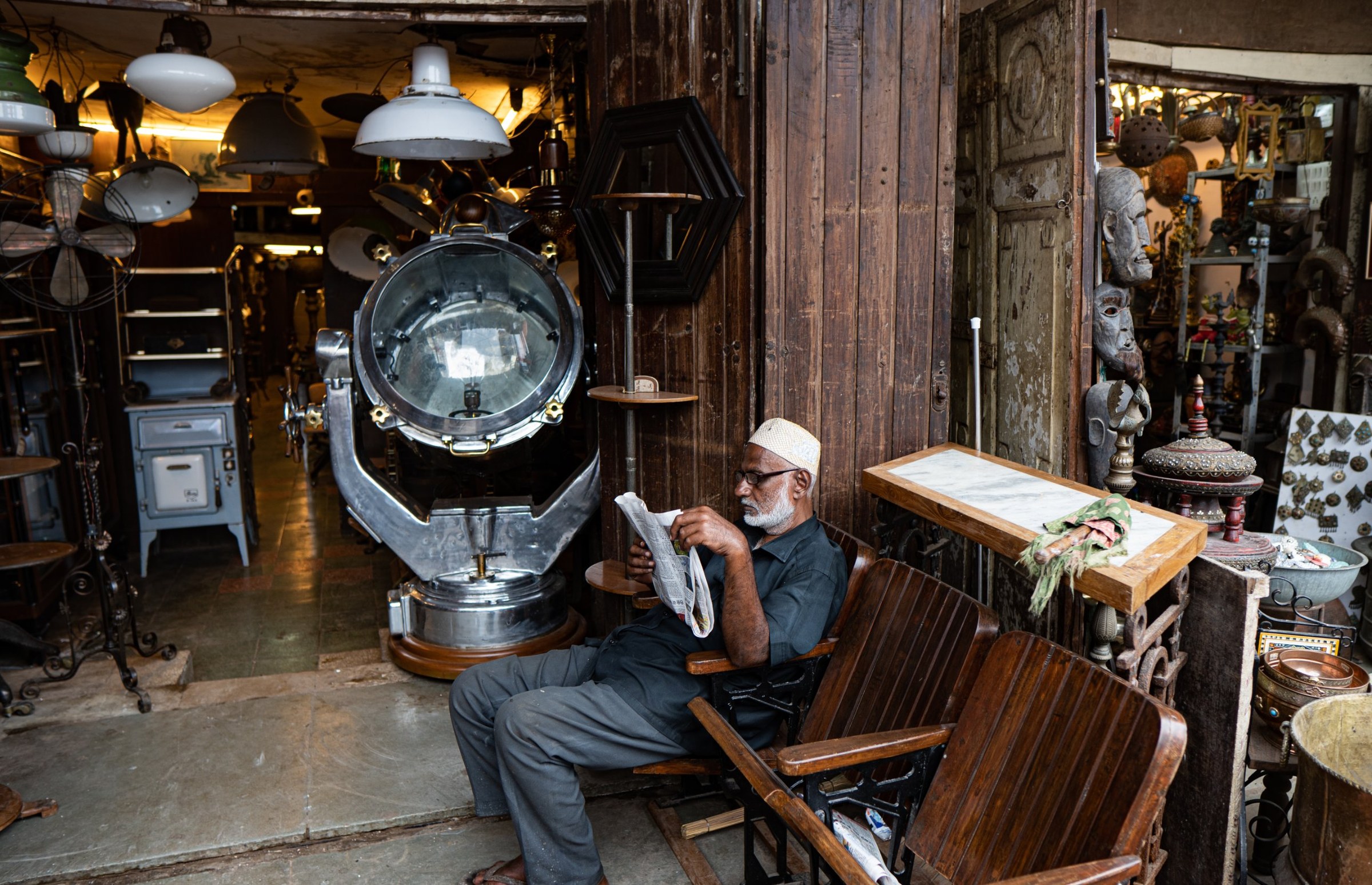 An elderly man reading a newspaper outside an antique shop, beside a large ship’s searchlight and hanging lamps