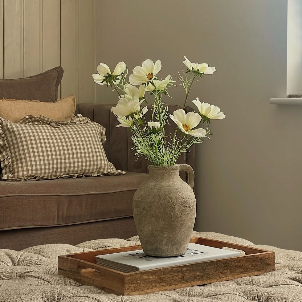 Rustic vase with white flowers on a wooden tray atop a cushioned ottoman. Brown sofa with checkered pillows in the cozy setting.