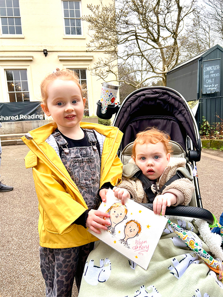 Child in yellow coat holding card next to baby in stroller.