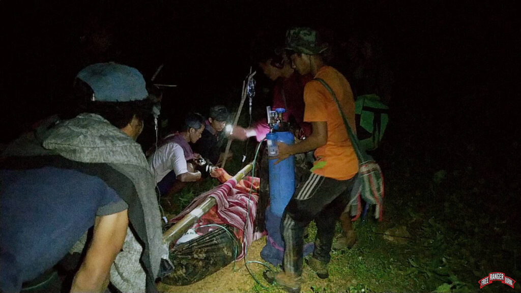 The Karenni Rangers provide medical care while moving the patient. He is being carried in a hammock attached to a bamboo pole.