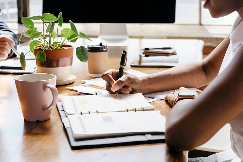 Person writing in a notebook at desk with coffee and potted plant.