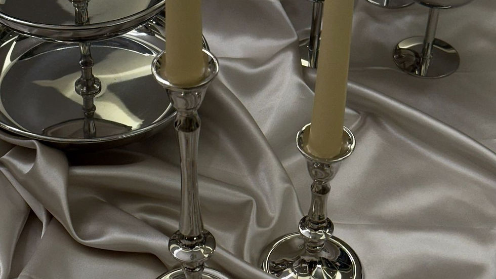 Silver candlesticks with cream candles on a silky draped fabric, surrounded by shiny silver dishes and bowls on a stone table.