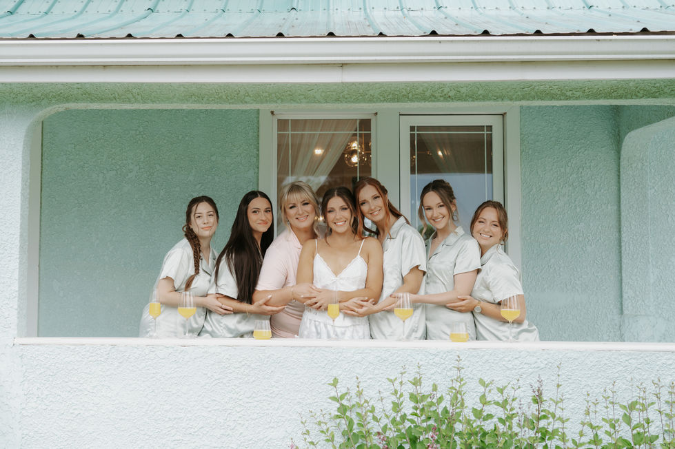 Bride and bridesmaids smiling together with drinks on the porch of the pale blue bridal house at River’s Edge Devon.