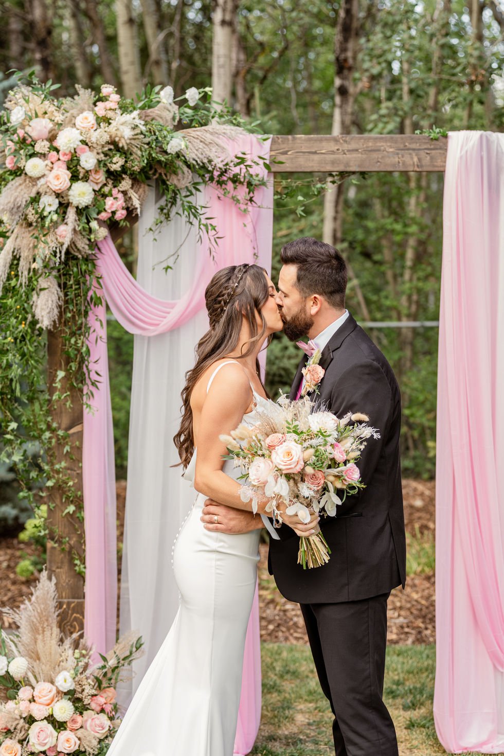Bride and groom kissing beneath a pink floral arch at their Edmonton backyard wedding ceremony.