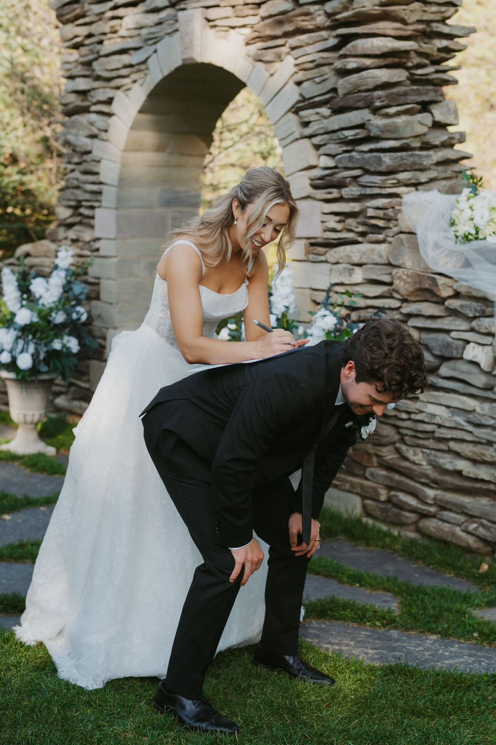 Bride playfully signing the registry on the groom’s back beside the stone ceremony arch at The Valley.
