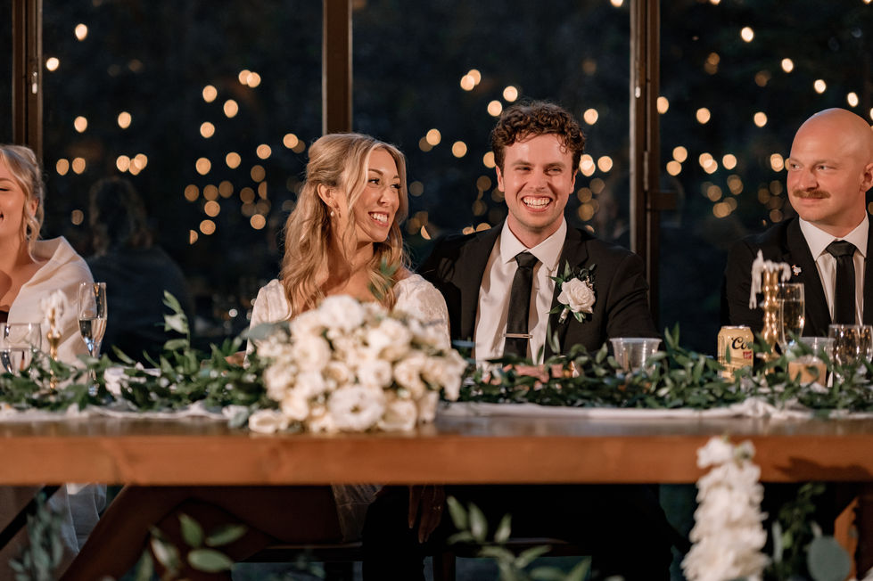 Bride and groom seated at a wooden head table with greenery and string lights at The Valley.